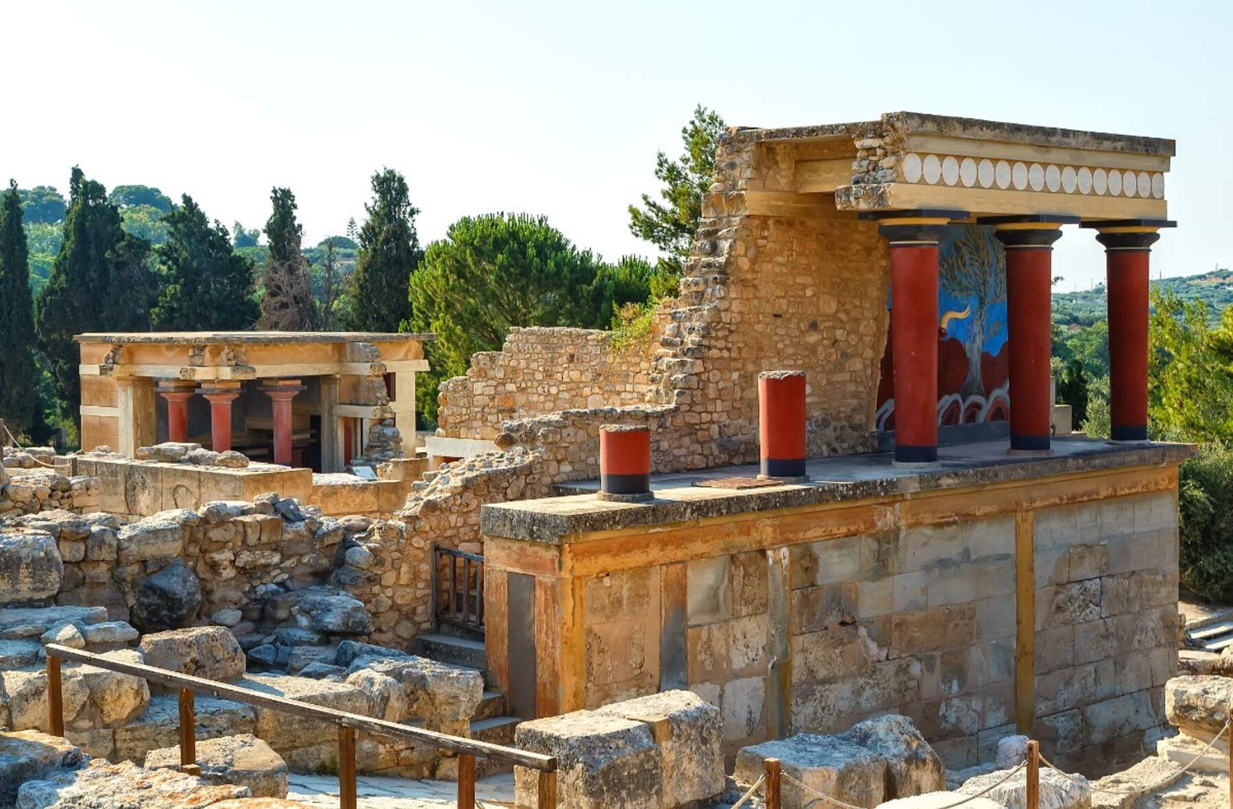 Ερείπια του Μινωικού Ανακτόρου της Κνωσού στην Κρήτη, με χαρακτηριστικές κόκκινες κολώνες και τοιχογραφίες στο φόντο.