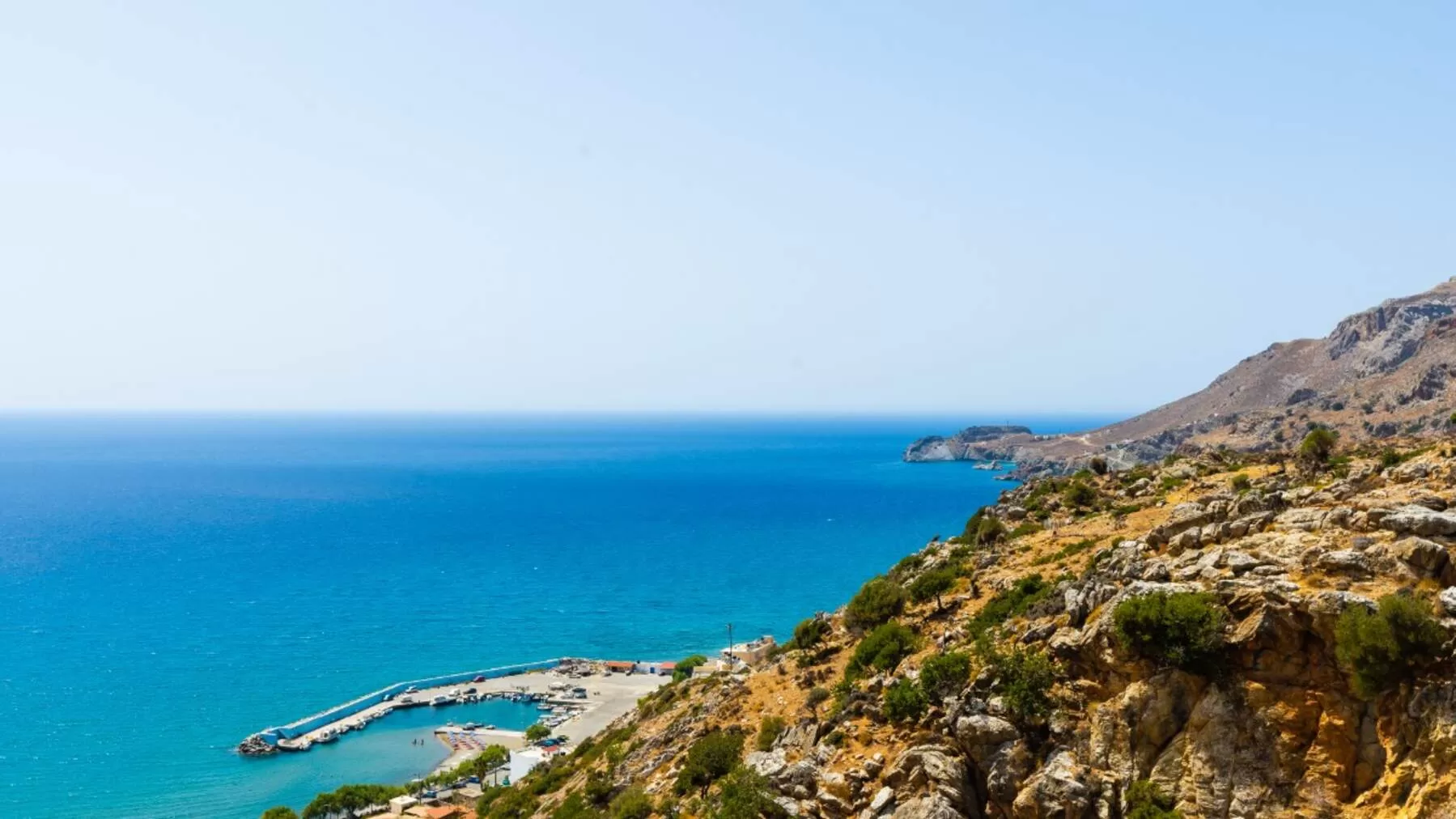 A scenic view of a rocky coastline with clear blue sea and a small harbor at the base of dry, shrub-covered hills under a bright sky.