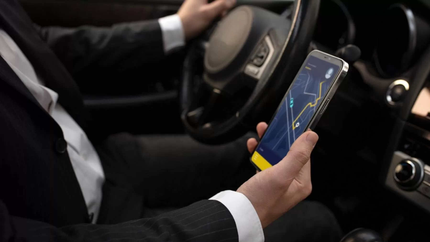 A man in a suit sitting in a car and holding a smartphone displaying a navigation or map application while driving.